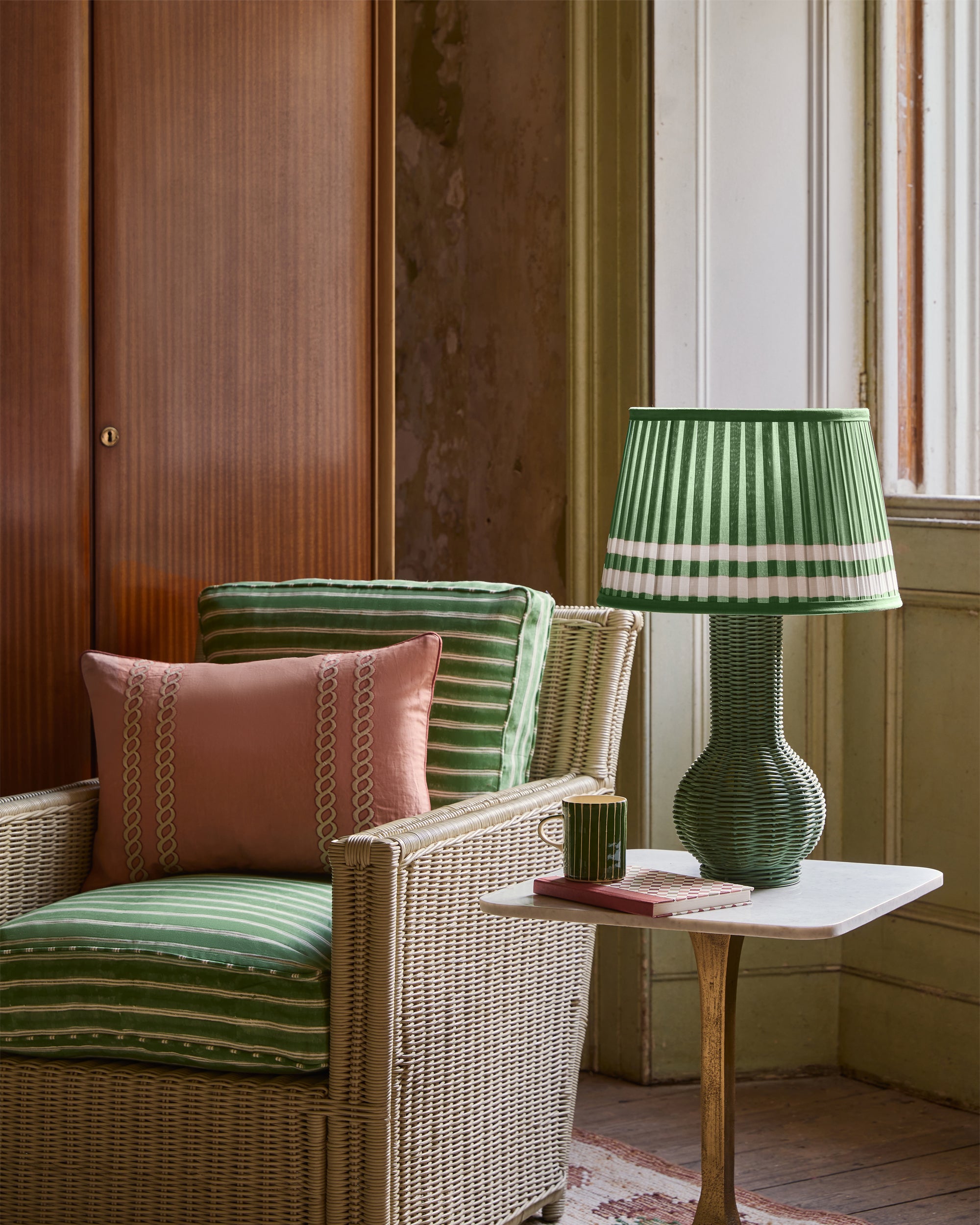 Wicker style chair with striped cushions and a green lamp in a room with wooden paneling.