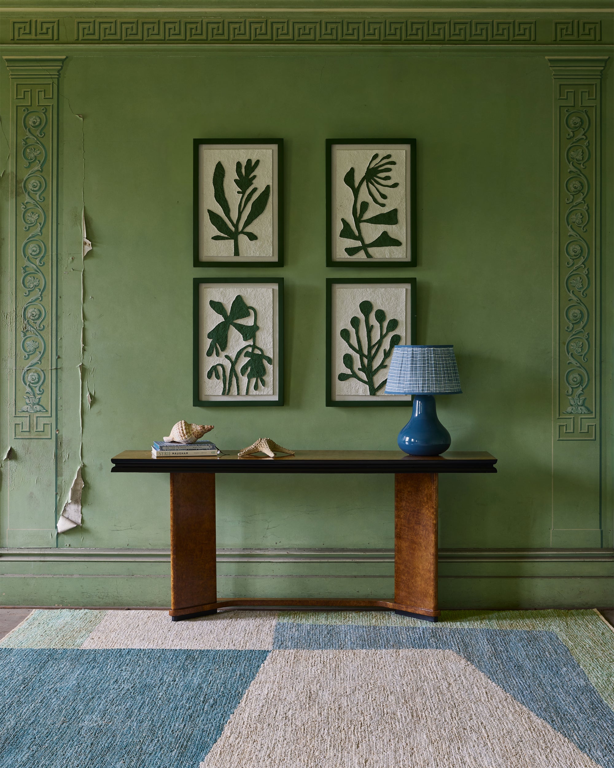 Green wall with framed botanical art and a wooden console table with a blue lamp.