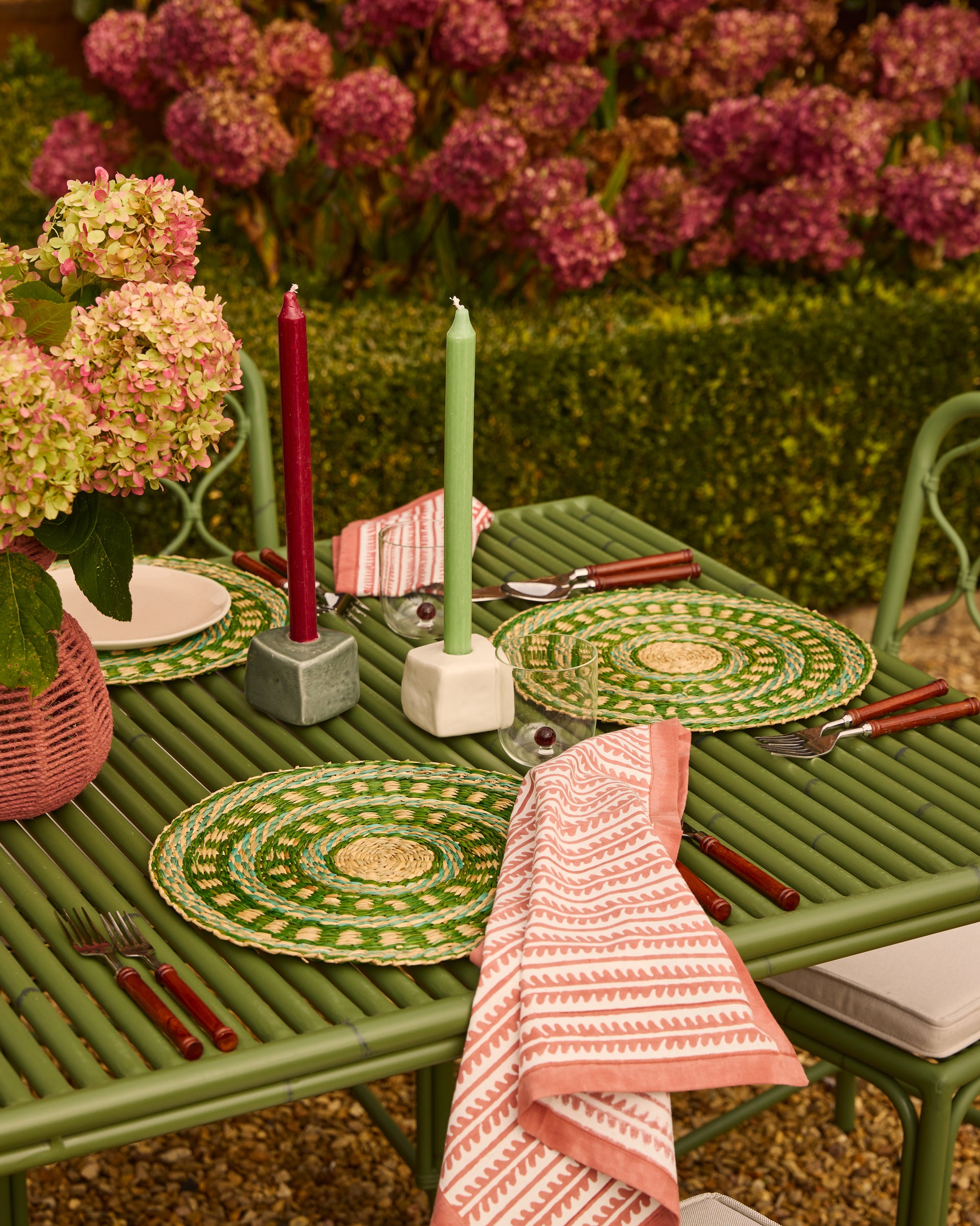 Outdoor table setting with green plates, red and green candles, and pink napkins on a green table.