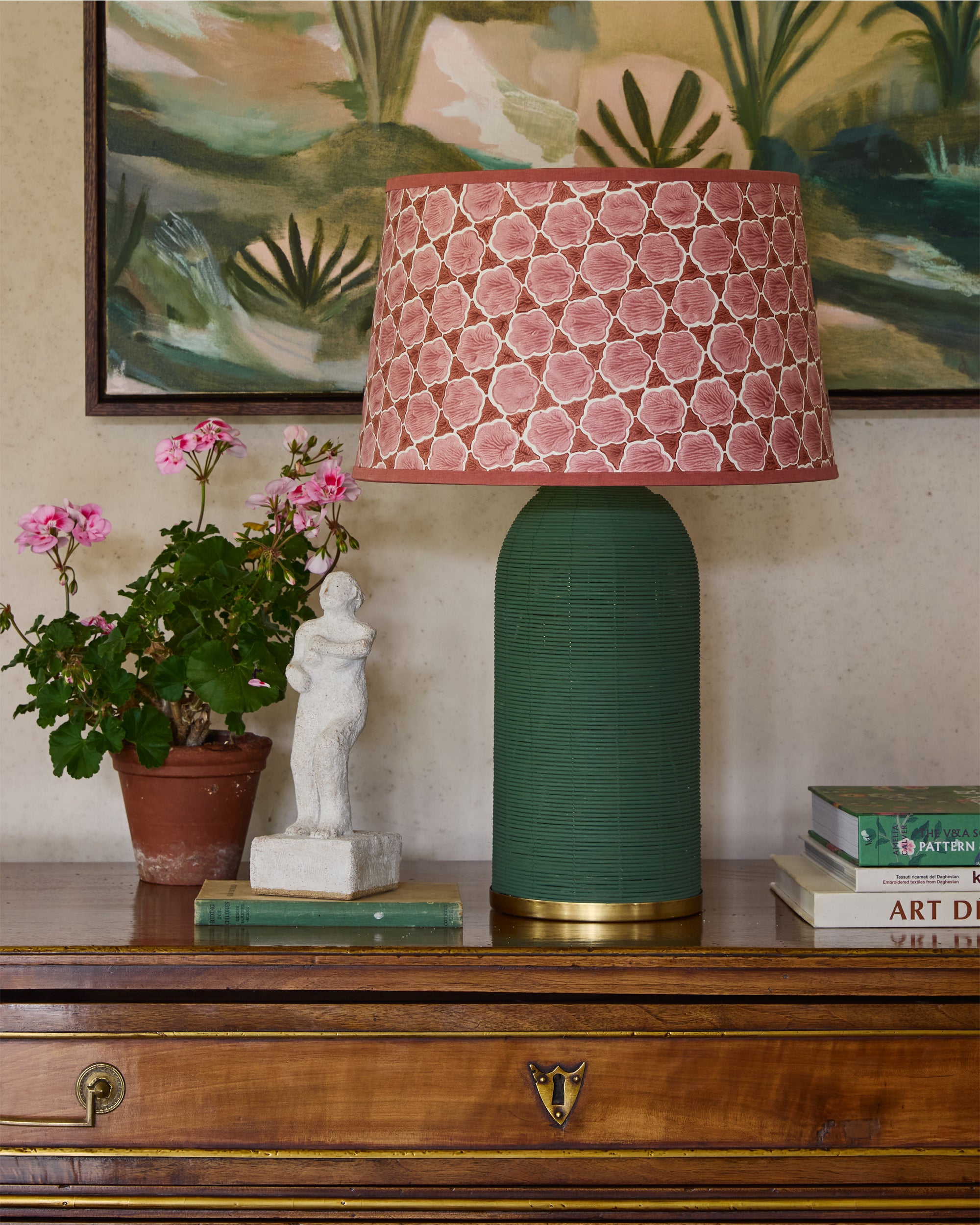 Decorative lamp with a pink patterned shade on a wooden surface with a plant and statue.