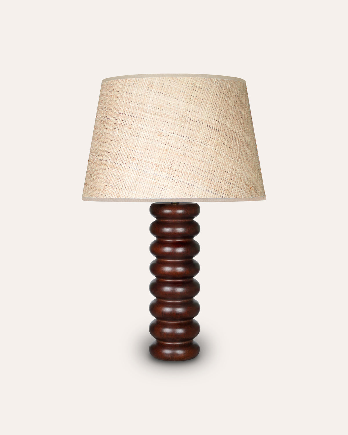 A table lamp with a seema raffia shade and a dark brown ribbed wooden base, shown against a solid off-white background.