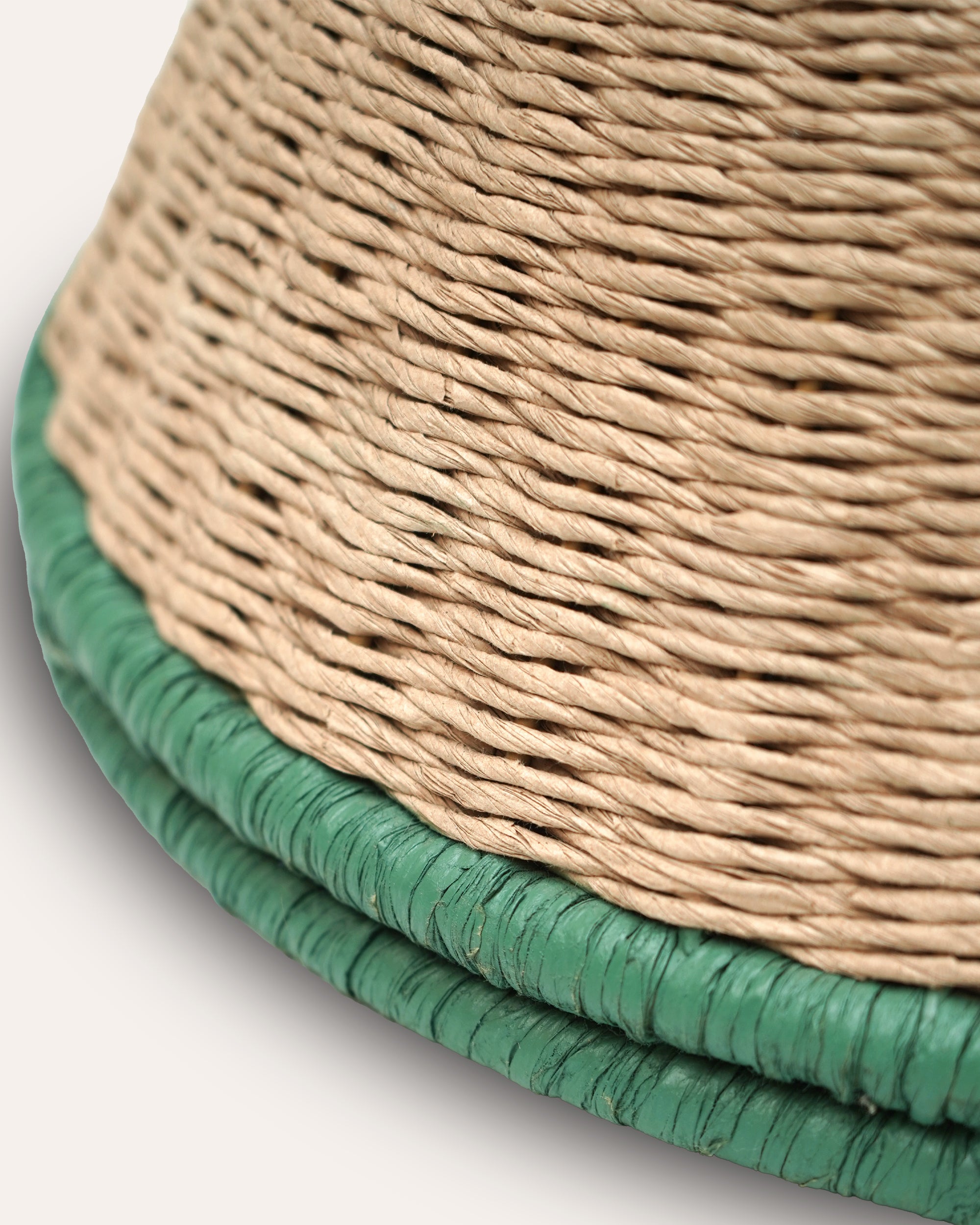 Close-up of a woven natural fiber pendant, showing a beige/tan body with a green braided trim along the lower edge on a light gray background.
